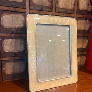 May include: A rectangular picture frame with a cream-coloured, marbled enamel border and a thin silver-toned metal edge. The frame features a smooth, glossy finish with subtle, swirling patterns throughout the border and a clear glass centre.