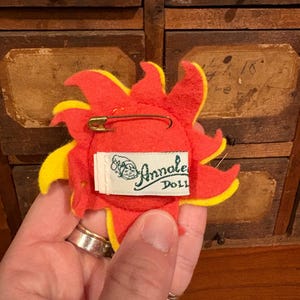 Puede incluir: Un broche de fieltro con forma de sol con detalles rojos y amarillos. La parte posterior del broche presenta un imperdible y una etiqueta blanca con el texto "Annalee Doll". El broche est&aacute; sostenido por una mano.