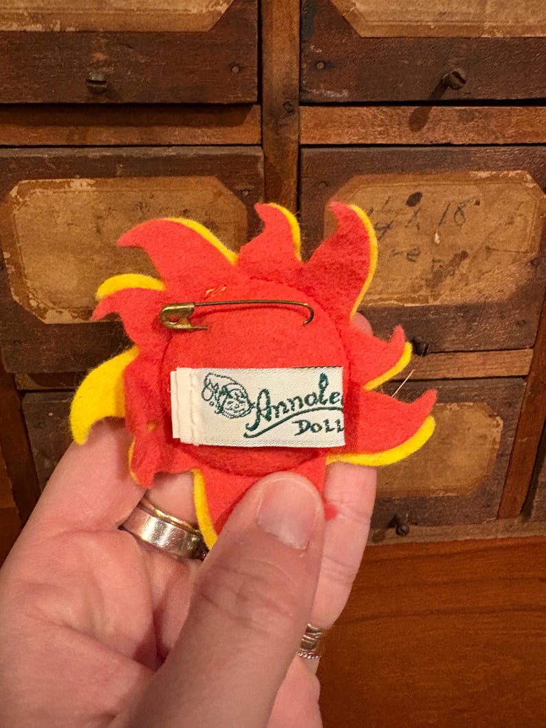 Puede incluir: Un broche de fieltro con forma de sol con detalles rojos y amarillos. La parte posterior del broche presenta un imperdible y una etiqueta blanca con el texto "Annalee Doll". El broche est&aacute; sostenido por una mano.