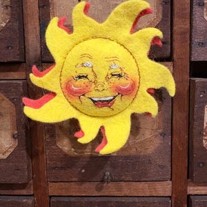 Puede incluir: Decoraci&oacute;n de sol de fieltro con una cara sonriente, cuerpo amarillo y detalles rojos. El sol tiene una expresi&oacute;n alegre con mejillas rosadas y probablemente est&aacute; hecho a mano. Es un objeto decorativo.
