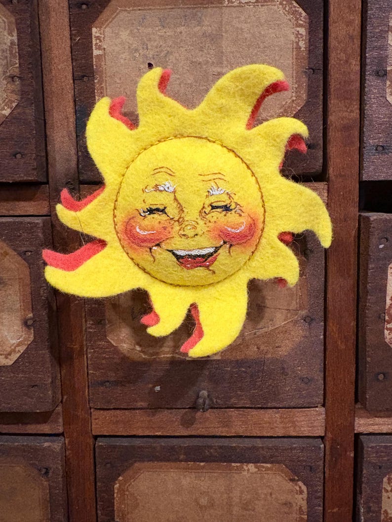 Puede incluir: Decoraci&oacute;n de sol de fieltro con una cara sonriente, cuerpo amarillo y detalles rojos. El sol tiene una expresi&oacute;n alegre con mejillas rosadas y probablemente est&aacute; hecho a mano. Es un objeto decorativo.