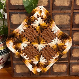 Op de afbeelding: Een gehaakt vierkant met een bruin, wit en geel madeliefjespatroon. Het vierkant is gemaakt met verschillende garenkleuren en -texturen.