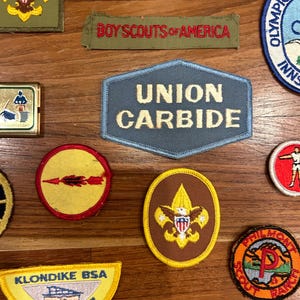 May include: A collection of vintage Boy Scouts of America patches, including a Union Carbide patch, a Klondike BSA patch, and a Philmont Scout Ranch patch. The patches are on a wooden surface.