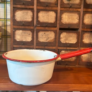 Puede incluir: Cacerola de esmalte blanco con asa roja. La cacerola tiene un borde rojo y un acabado de esmalte desgastado.