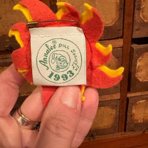 Puede incluir: Un broche de fieltro con forma de flor en tonos rojos y amarillos, con un imperdible en la parte posterior. El broche tiene una etiqueta que dice "Annalee Doll Society 1993". Los p&eacute;talos de la flor son superpuestos y detallados.