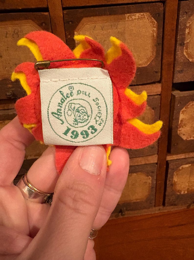 Puede incluir: Un broche de fieltro con forma de flor en tonos rojos y amarillos, con un imperdible en la parte posterior. El broche tiene una etiqueta que dice "Annalee Doll Society 1993". Los p&eacute;talos de la flor son superpuestos y detallados.