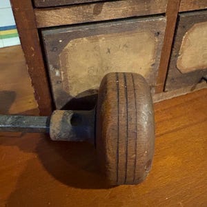 Puede incluir: Un pomo de puerta de madera vintage con un acabado redondo de color marrón oscuro y tres ranuras horizontales alrededor de su circunferencia. El pomo está unido a un eje metálico cuadrado y desgastado que se extiende hacia la izquierda.