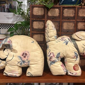May include: Two vintage fabric stuffed animal toys. A light brown elephant with floral print and a light brown dog with floral print. The toys are sitting on a wooden shelf.