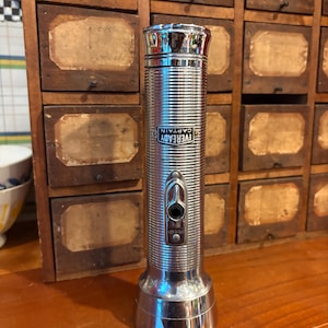 May include: A vintage chrome flashlight with the text "Eveready Captain" on the side. The flashlight has a ribbed body, a push-button switch, and a reflective top and base. It is standing on a wooden surface.