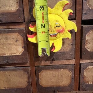 Puede incluir: Un sol de fieltro amarillo con una cara sonriente y detalles rojos, que mide aproximadamente 7,6 cm de altura. El sol est&aacute; colocado sobre un mueble de madera vintage con varios cajones, cada uno con una etiqueta.
