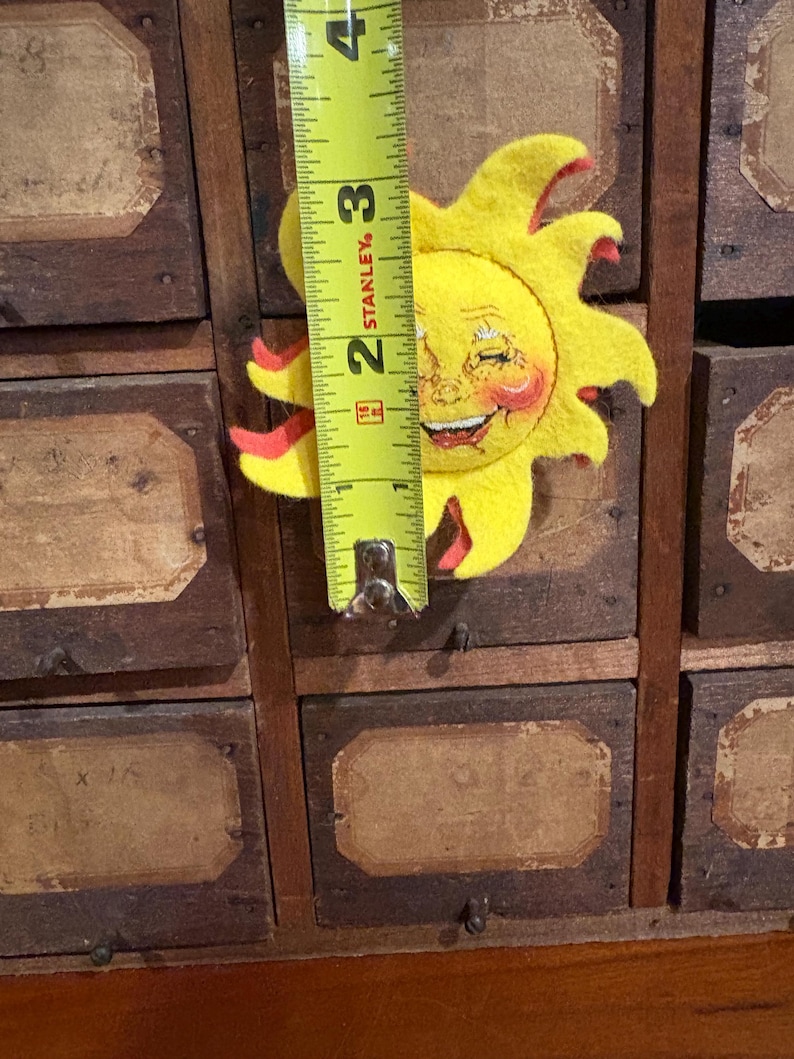 Puede incluir: Un sol de fieltro amarillo con una cara sonriente y detalles rojos, que mide aproximadamente 7,6 cm de altura. El sol est&aacute; colocado sobre un mueble de madera vintage con varios cajones, cada uno con una etiqueta.