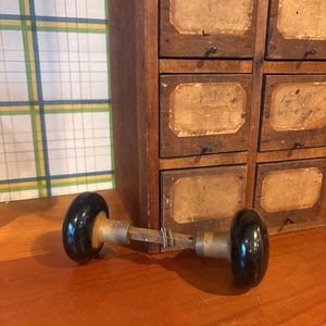 May include: A vintage door knob set featuring two round, glossy black knobs connected by a central metal spindle. The hardware includes brass-toned collars at the base of each knob, with a small metal wire wrapped around the square connecting rod.