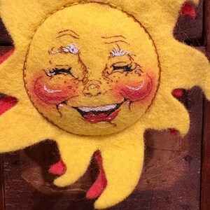 Puede incluir: Decoraci&oacute;n artesanal de sol sonriente. El sol es principalmente amarillo con acentos rojos en los rayos y las mejillas. La cara presenta ojos cerrados, una boca roja sonriente y mejillas rosadas, creando una est&eacute;tica alegre.