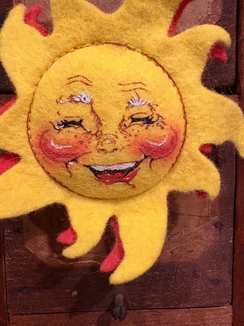 Puede incluir: Decoraci&oacute;n artesanal de sol sonriente. El sol es principalmente amarillo con acentos rojos en los rayos y las mejillas. La cara presenta ojos cerrados, una boca roja sonriente y mejillas rosadas, creando una est&eacute;tica alegre.