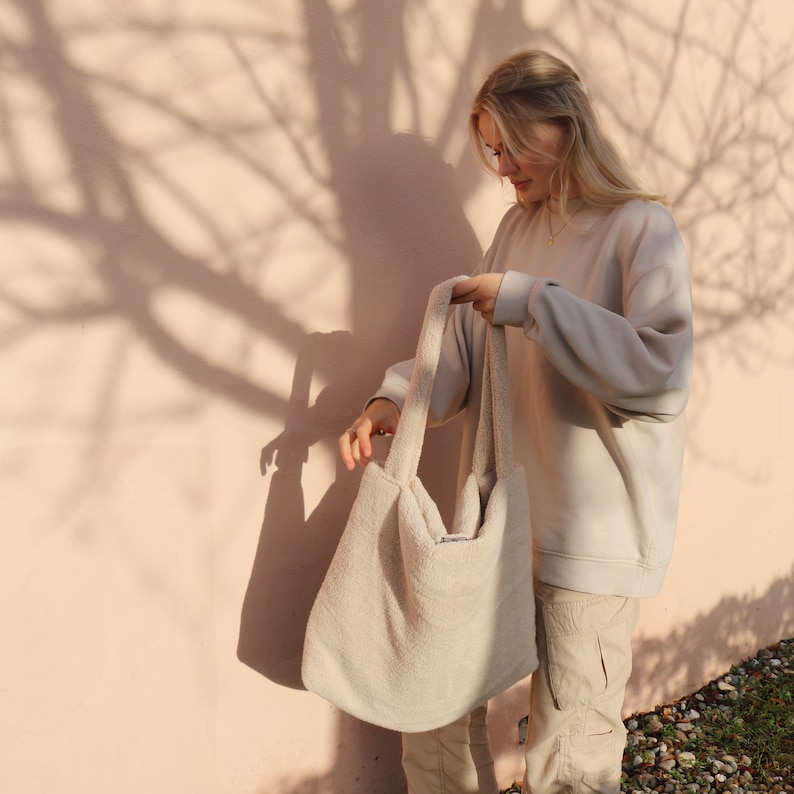 Puede incluir: Una mujer sostiene una gran bolsa de mano de color blanco roto y esponjosa. La bolsa tiene una textura suave y peluda y est&aacute; hecha de una tela gruesa y tejida. La bolsa es lo suficientemente grande como para llevar una variedad de art&iacute;culos.
