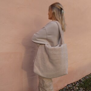 Puede incluir: Un bolso tote grande, esponjoso, de color blanco roto con una correa larga. El bolso est&aacute; hecho de un material suave y afelpado y lo lleva una persona que lleva un su&eacute;ter beige claro.