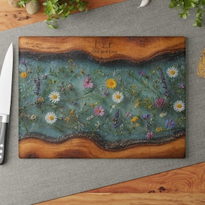 May include: A rectangular wooden cutting board with a turquoise resin center filled with colorful wildflowers. The board has a natural wood grain border and the names "John and Elsa" are engraved on the top.