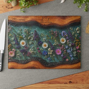 May include: Wooden rectangular chopping board with a teal resin centre, inlaid with colourful dried flowers. The board has a natural wood grain border and the text "J & E" with a heart and "John and Elsa" is engraved on the top.