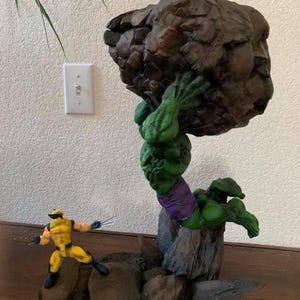 May include: A green Hulk figure lifting a large brown rock, with a yellow Wolverine figure in front. The Hulk is wearing purple shorts. The figures are on a rocky base.