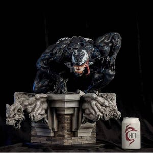 May include: A detailed statue of the Marvel Comics character Venom, in a crouching pose, atop a stone base with gargoyle figures. The statue is predominantly black with white accents and a red tongue. A white can with red text is to the right.