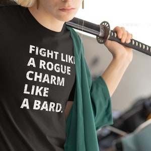May include: A person wearing a black t-shirt with the text "FIGHT LIKE A ROGUE CHARM LIKE A BARD." The person is holding a black sword over their shoulder. The person is wearing a green jacket and sunglasses.