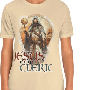 Puede incluir: Una camiseta beige con un gr&aacute;fico de un hombre con una t&uacute;nica sosteniendo un bast&oacute;n y un globo terr&aacute;queo. El texto "JESUS is my CLERIC" est&aacute; impreso debajo del gr&aacute;fico.