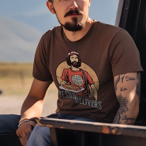 May include: A man wearing a brown t-shirt with a graphic of a person holding a pizza and the text "Jesus Delivers". He has a beard and tattoos on his arm.