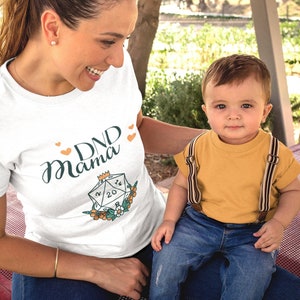 May include: A woman wearing a white t-shirt with the text "DND Mama" and a graphic of a twenty-sided die. She is sitting on a red bench with a young child sitting on her lap. The child is wearing a yellow shirt and blue jeans.