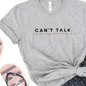 K&ouml;nnte beinhalten: Ein graues T-Shirt mit dem Text "CAN'T TALK I'm thinking about gaming." in schwarzer und ros&eacute;goldener Schrift. Das Shirt wird mit Jeans-Shorts und pinkfarbenen und blauen Flip-Flops kombiniert.