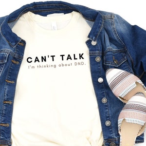May include: A white t-shirt with the text "CAN'T TALK I'm thinking about DND." printed in black.