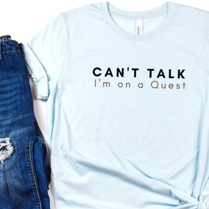 May include: A light blue t-shirt with the text "CAN'T TALK I'm on a Quest" in black and brown lettering.