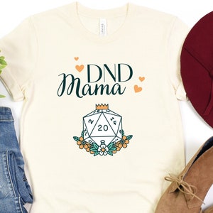 May include: A white t-shirt with the text "DND Mama" in green and black lettering. A stylized illustration of a twenty-sided die with a crown on top is below the text. The die is surrounded by orange and white flowers.