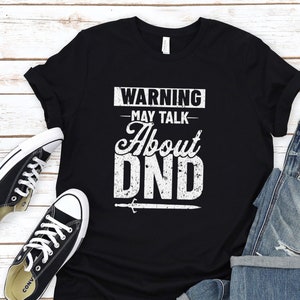May include: Black t-shirt with white text that reads "WARNING - MAY TALK ABOUT DND" with a sword graphic. The shirt is on a white wood background with a pair of black sneakers and blue jeans.
