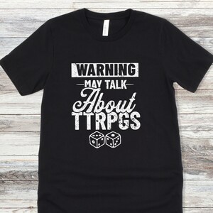 May include: Black t-shirt with white text that reads "WARNING - MAY TALK ABOUT TTRPGS" and two white dice.