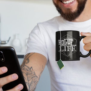 Op de afbeelding: Een persoon houdt een zwarte mok vast met de tekst "Rogue Life" erop gedrukt. De mok is gevormd als een 20-zijdige dobbelsteen. De persoon glimlacht en kijkt naar een smartphone.