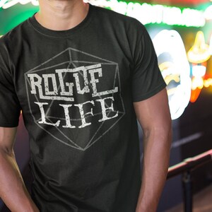 May include: A black t-shirt with a white graphic of a 20-sided die with the text "Rogue Life" inside. The person wearing the shirt is standing in front of a neon sign that says "Frontier Whiskey".