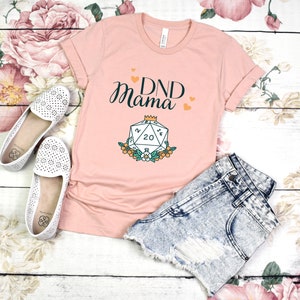 May include: A pink t-shirt with the text "DND Mama" and a graphic of a twenty-sided die with a crown on top. The die is surrounded by flowers.
