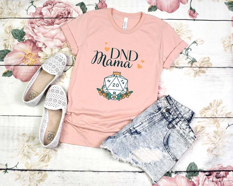 May include: A pink t-shirt with the text "DND Mama" and a graphic of a twenty-sided die with a crown on top. The die is surrounded by flowers.