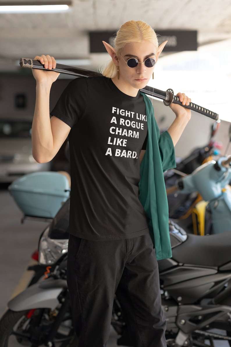 May include: A person wearing a black t-shirt with the text "FIGHT LIKE A ROGUE CHARM LIKE A BARD." The person is holding a black sword over their shoulder. The person is wearing a green jacket and sunglasses.