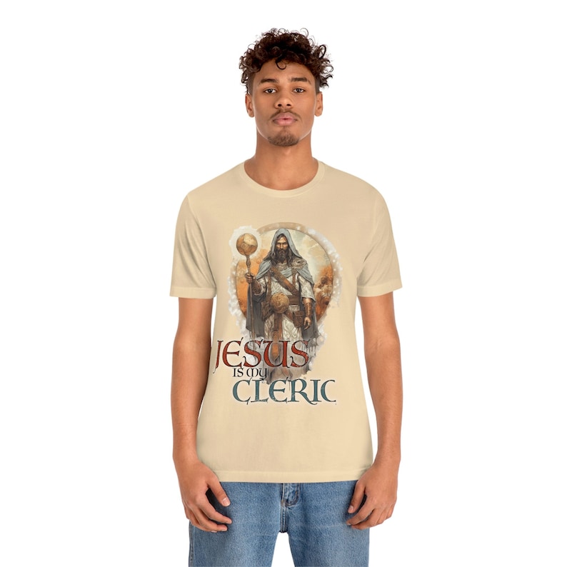 Puede incluir: Una camiseta beige con un gr&aacute;fico de un hombre con una t&uacute;nica sosteniendo un bast&oacute;n y un globo terr&aacute;queo. El texto "JESUS is my CLERIC" est&aacute; impreso debajo del gr&aacute;fico.