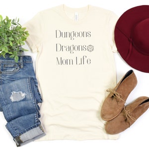 May include: A cream colored t-shirt with the text "Dungeons Dragons Mom Life" and a small graphic of a 20-sided die.