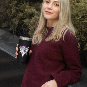 K&ouml;nnte beinhalten: Ein schwarzer Edelstahlbecher mit einer wei&szlig;en Grafik, auf der "Stranger Things" in einem rosa Dreieck steht. Der Becher wird von einer Person gehalten, die einen kastanienbraunen Pullover tr&auml;gt.