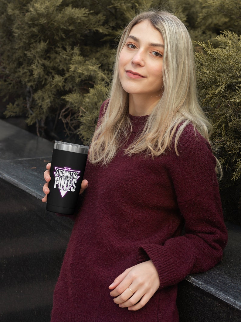 K&ouml;nnte beinhalten: Ein schwarzer Edelstahlbecher mit einer wei&szlig;en Grafik, auf der "Stranger Things" in einem rosa Dreieck steht. Der Becher wird von einer Person gehalten, die einen kastanienbraunen Pullover tr&auml;gt.