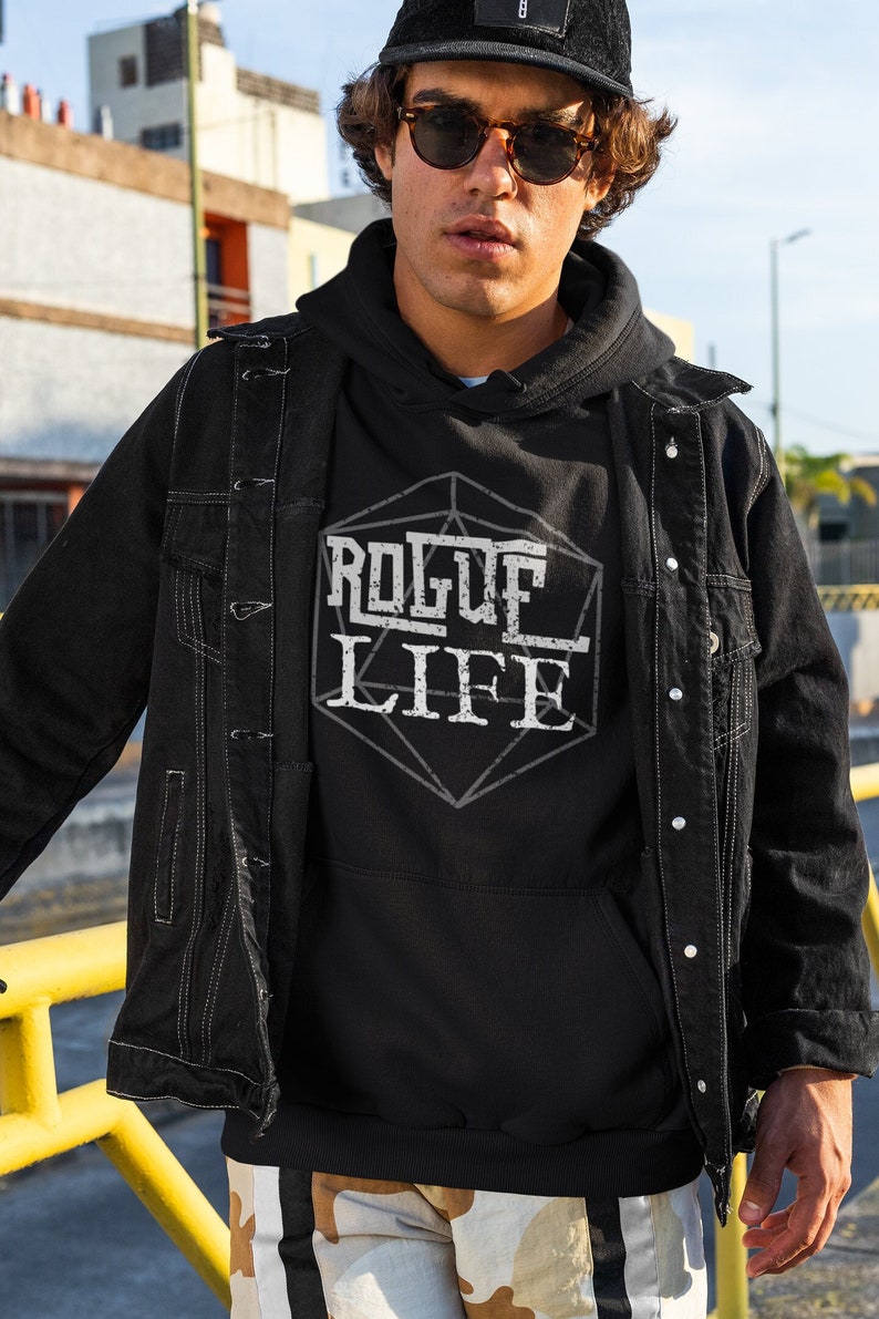 May include: A black hoodie with a white graphic of a 20-sided die with the text "ROGUE LIFE" inside. The person wearing the hoodie is wearing a black denim jacket and a black baseball cap.
