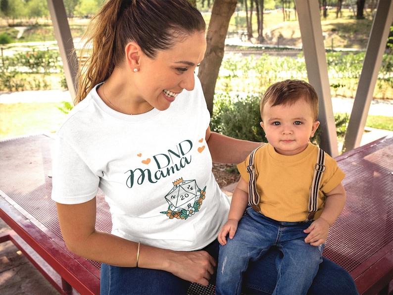 May include: A woman wearing a white t-shirt with the text "DND Mama" and a graphic of a twenty-sided die. She is sitting on a red bench with a young child sitting on her lap. The child is wearing a yellow shirt and blue jeans.