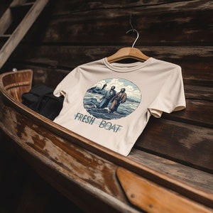 K&ouml;nnte beinhalten: Ein beiges T-Shirt mit einem Aufdruck von zwei Personen, die auf dem Wasser gehen, mit einem Leuchtturm im Hintergrund. Der Text "FRESH OFF THE BOAT" ist unter dem Aufdruck gedruckt.
