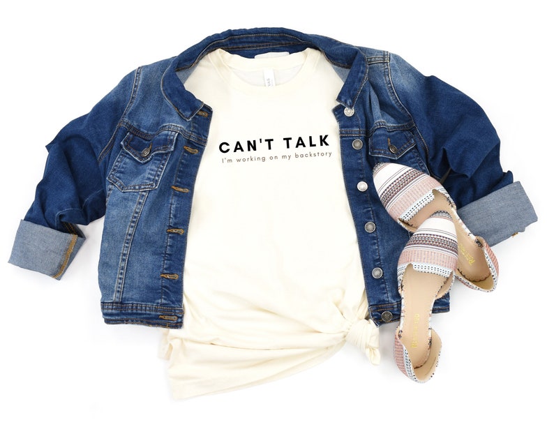 May include: A white t-shirt with the text "CAN'T TALK I'm working on my backstory" printed on it. The shirt is paired with a blue denim jacket and striped flats.