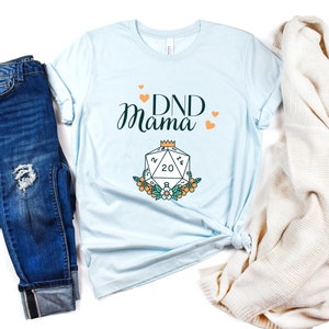 May include: A light blue t-shirt with the text "DND Mama" in black script. A stylized illustration of a twenty-sided die with a crown on top is below the text. The die is surrounded by flowers.
