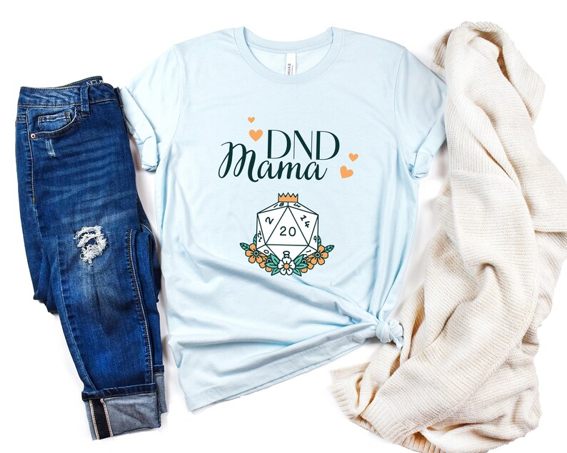 May include: A light blue t-shirt with the text "DND Mama" in black script. A stylized illustration of a twenty-sided die with a crown on top is below the text. The die is surrounded by flowers.