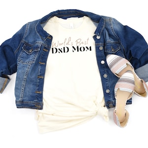 May include: A white t-shirt with the text "World's Best DND Mom" in black and brown lettering. The shirt is paired with a denim jacket and striped flats.
